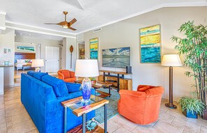 A spacious living room with a blue sofa, orange armchair, and a coffee table. There is a television on a wooden stand, a floor lamp, and a large painting on the wall.