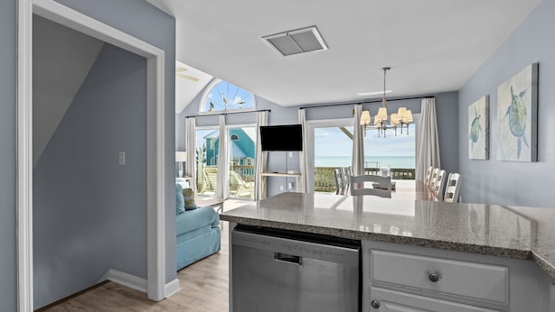 A modern kitchen with a dishwasher, a dining area with a view of the ocean, and a living room with a blue sofa. - Bright, spacious oceanfront duplex with pool access (Emerald Isle)