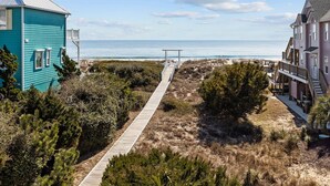 A coastal property with a wooden walkway leading to a beach, surrounded by dense vegetation and two buildings, one blue and one pink. - Bright, spacious oceanfront duplex with pool access (Emerald Isle)