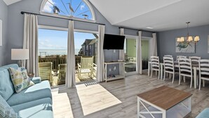 A spacious living room with a large window, a flat-screen TV, a sofa, and a dining area with chairs. - Bright, spacious oceanfront duplex with pool access (Emerald Isle)