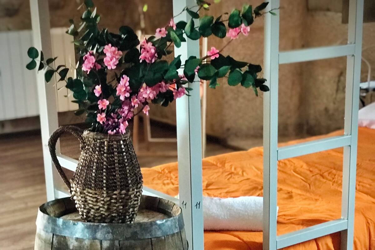 Ein rustikaler Holzbottich als Pflanzgefäß mit rosa Blüten, vor einem weißen Metallbettgestell mit orangefarbener Bettdecke platziert.
