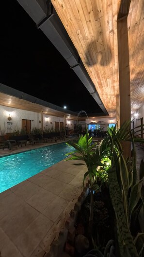 Une piscine intérieure avec un plafond en bois et un sol carrelé, entourée de chaises longues et de plantes en pot.