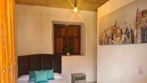 A bedroom with a bed, a nightstand, a window with a plant, and a cityscape painting on the wall. - Hotel Alcazar Real (Útica)