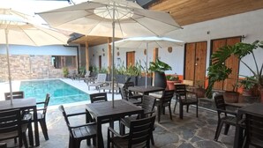 An outdoor patio area with stone flooring, wooden tables and chairs, umbrellas, and a pool in the background. - Hotel Alcazar Real (Útica)