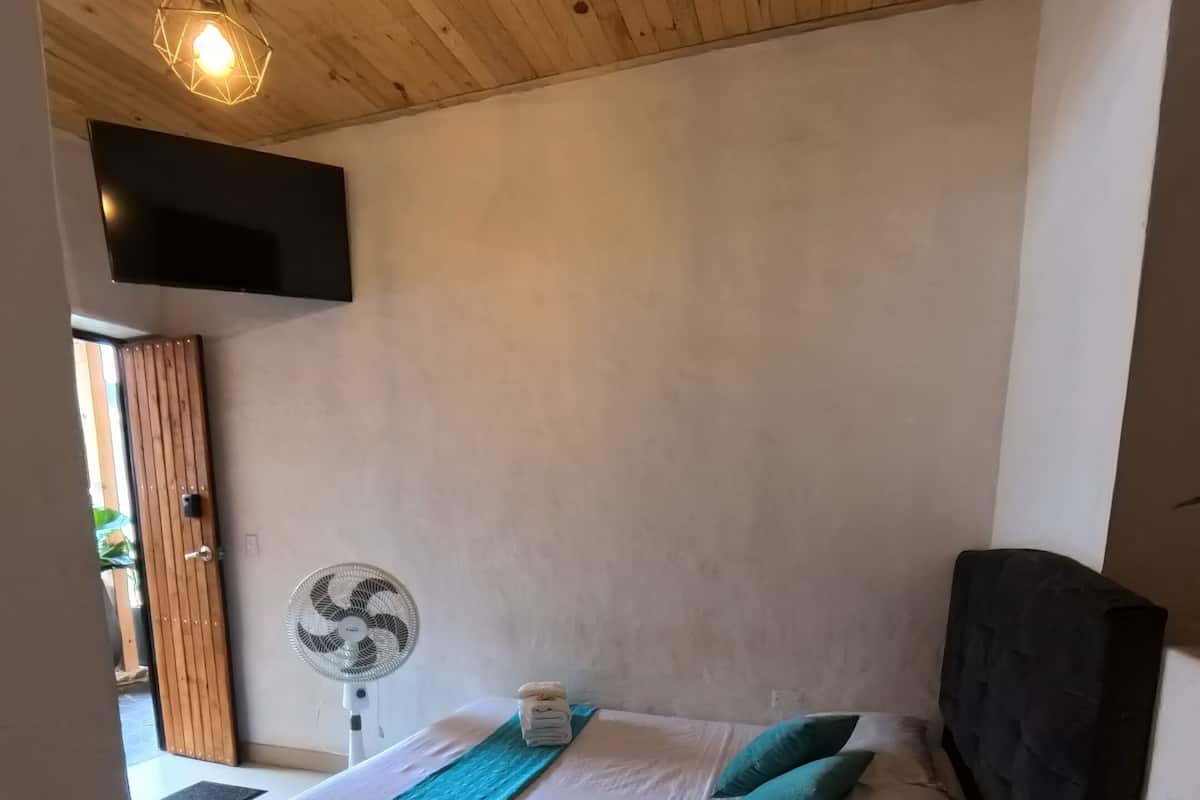 Ein Schlafzimmer mit Holzboden, einem Bett mit schwarzem Kopfteil, einem Nachttisch, einem Ventilator und einem an der Wand befestigten Fernseher.
