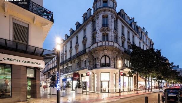 Ein historisches Gebäude mit aufwendiger Architektur, ein Laden mit der Beschilderung 'CREDIT AGRICOLE' und eine Straße mit Autos und Bäumen.
