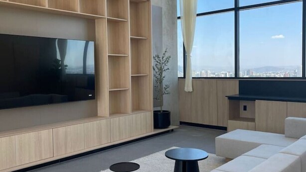 A modern living room with a large flat-screen TV, built-in wooden shelving, a large window with a city view, and a sectional sofa. - Great New Modern Department Expo GDL (Guadalajara)