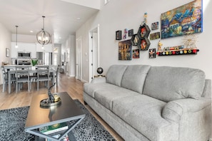 A modern living room with a grey sofa, a dining area with a white table and chairs, and a kitchen in the background.