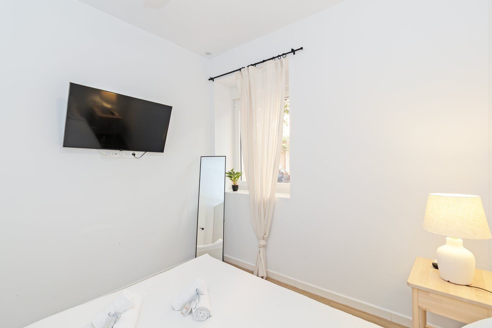 Une salle de bain moderne avec un grand miroir, un téléviseur à écran plat fixé au mur, une baignoire, une table de chevet avec une lampe et une fenêtre avec des rideaux.