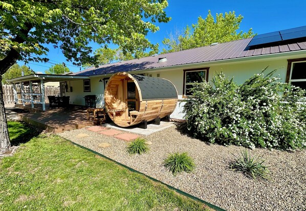 A house with a large wooden barrel-shaped structure attached to the front, a covered porch, and a garden with various plants.