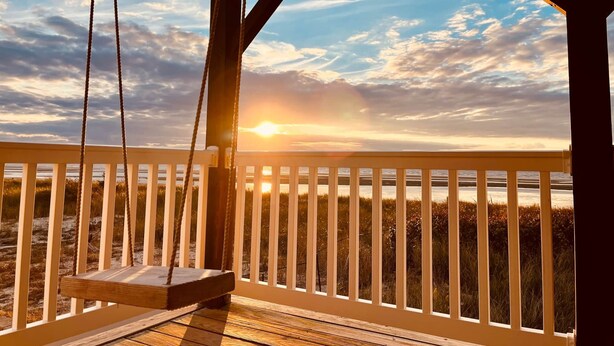 Una terraza de madera con un columpio frente a una playa al atardecer.