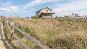 Una casa frente a la playa con balcón, rodeada de hierba alta y una valla de madera.