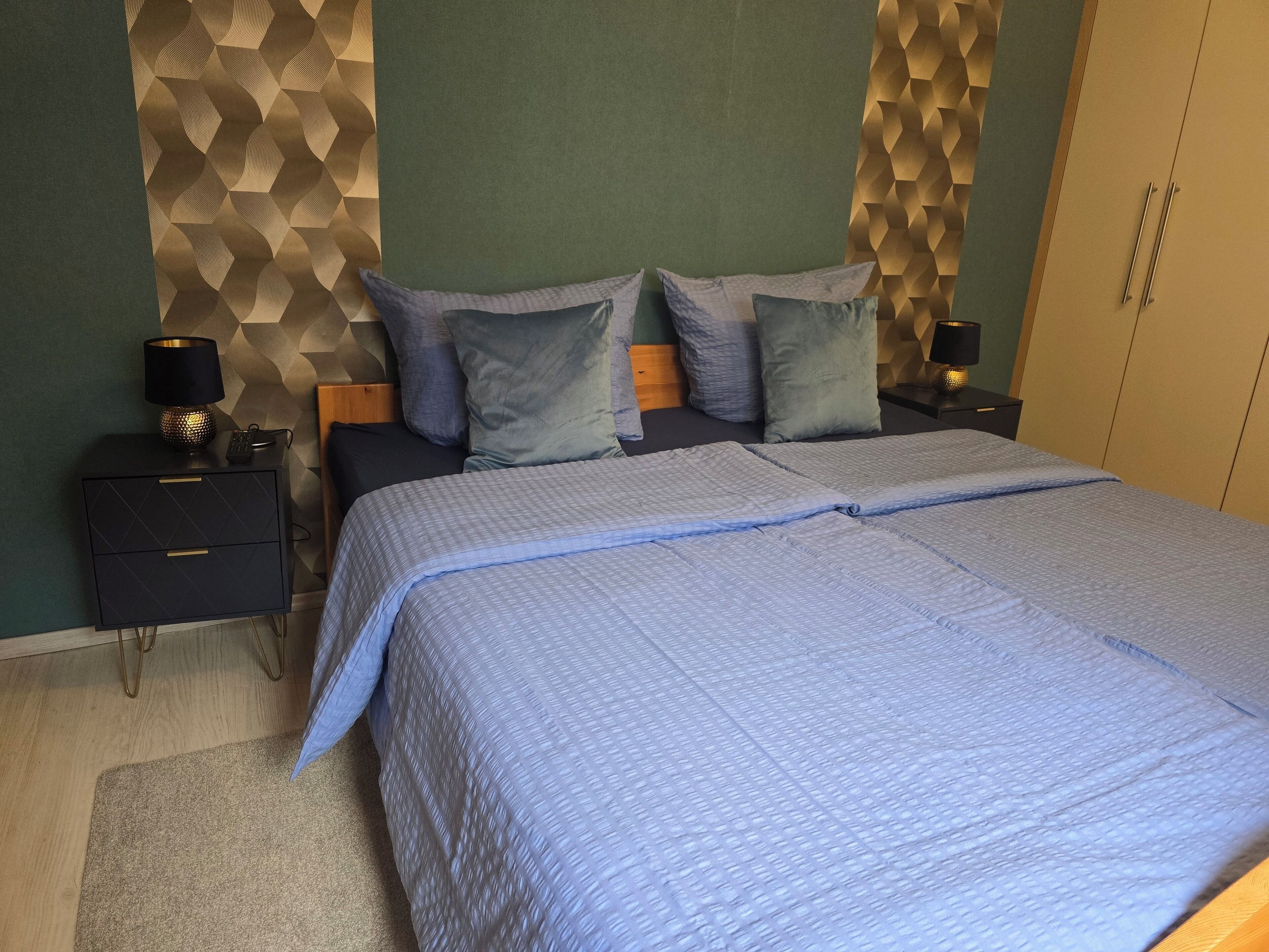 A neatly made bed with a blue quilt, two bedside tables with lamps, and a geometric-patterned wall.