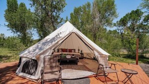 A dome-shaped tent with a bed, two chairs, and a small table, set on a wooden deck surrounded by trees.