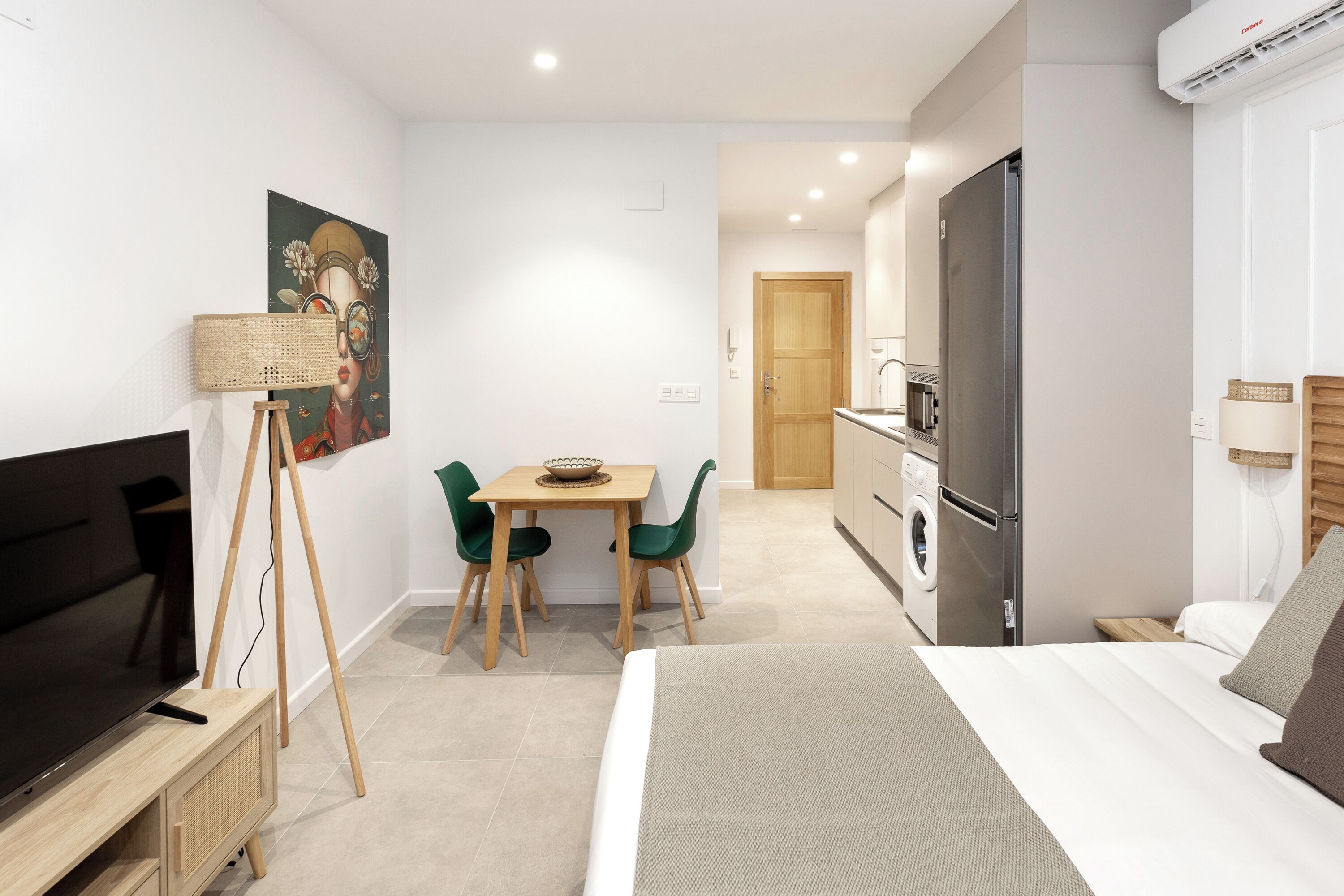 Una habitación de hotel moderna con cama, televisor, zona de comedor con sillas, cocina con refrigerador y lavadora.