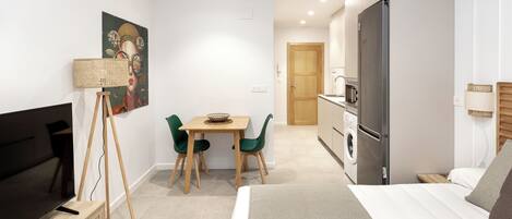 Una habitación de hotel moderna con cama, televisor, zona de comedor con sillas, cocina con refrigerador y lavadora.