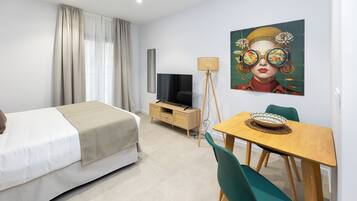 A modern hotel room with a bed, a TV, a dining table with chairs, and a painting on the wall.