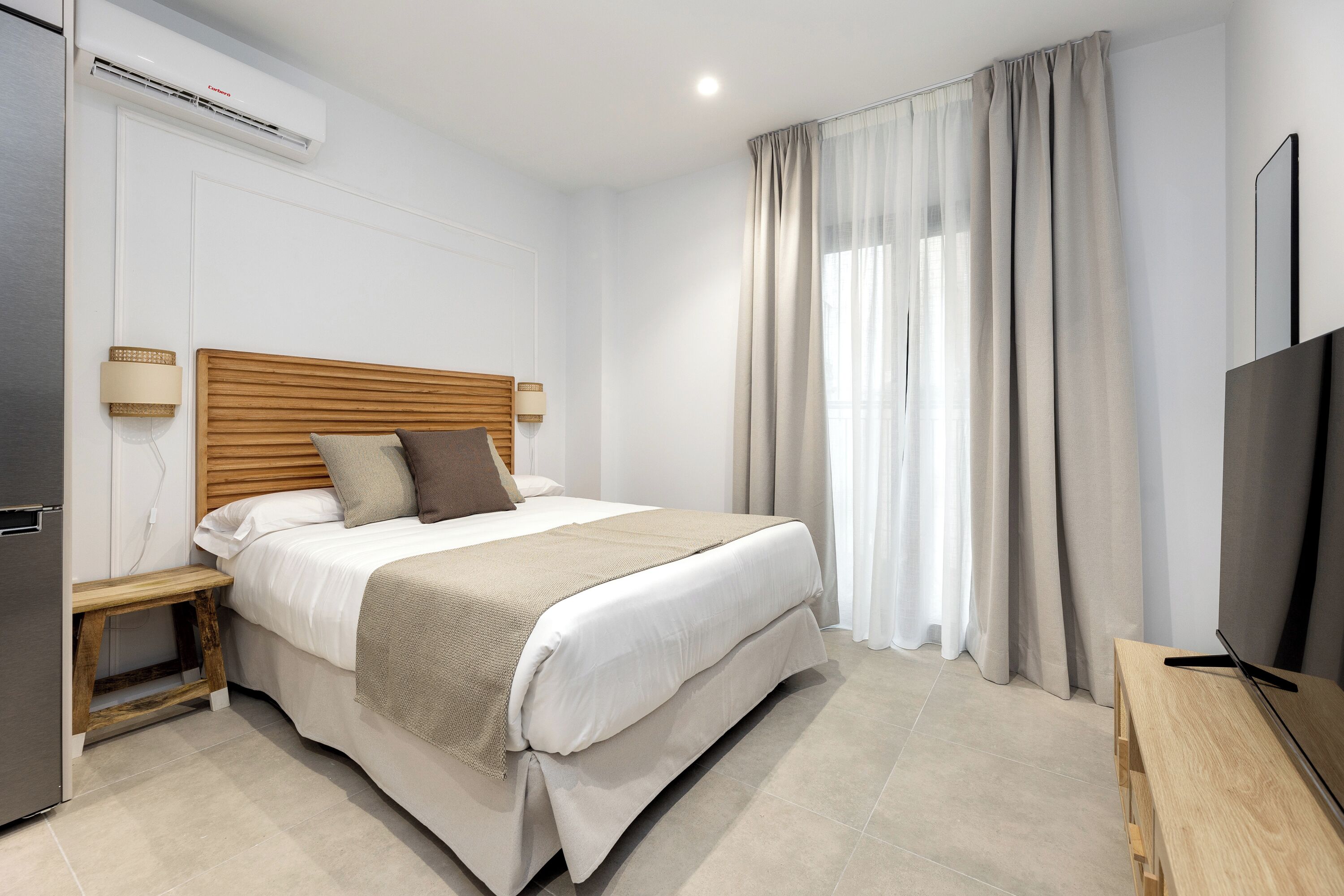 A hotel room with a bed, bedside table, lamp, air conditioning unit, and a TV.