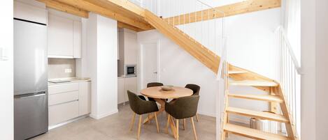 Una cocina moderna con armarios blancos, un refrigerador de acero inoxidable y un comedor con una mesa redonda y cuatro sillas. Una escalera de madera conduce a un nivel superior.