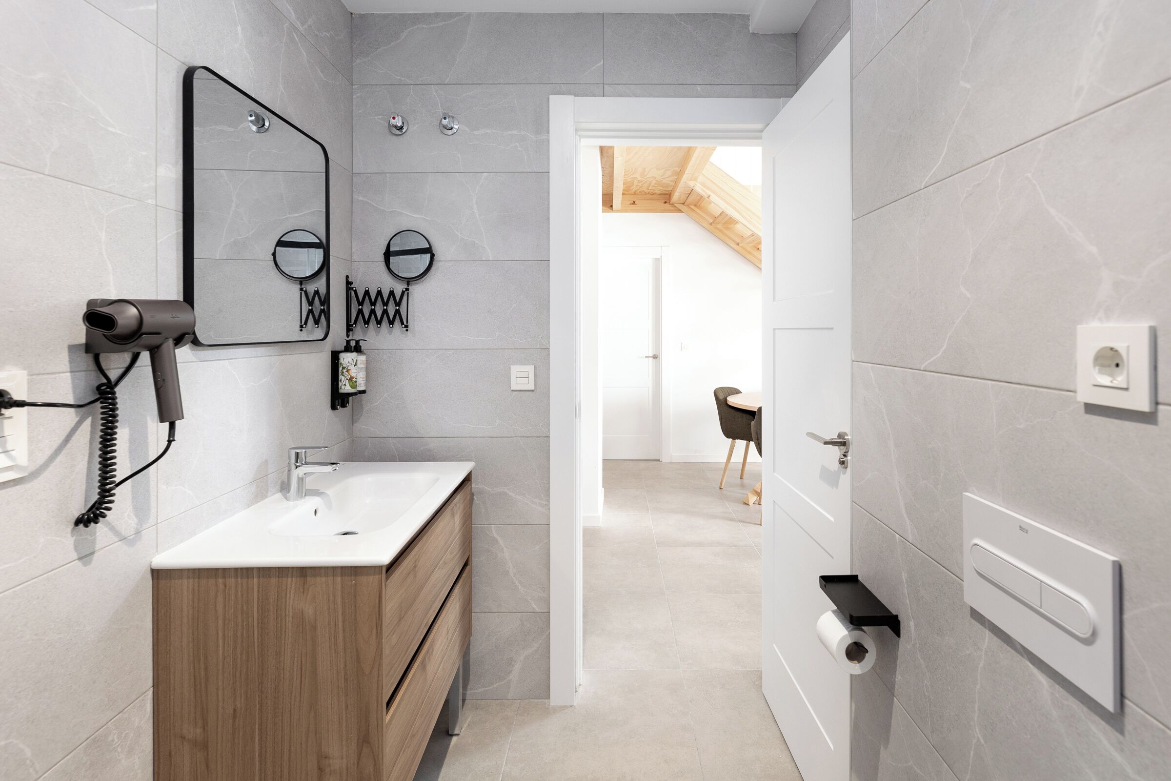 A modern bathroom with a wooden vanity, a large mirror, and a hairdryer on the wall.