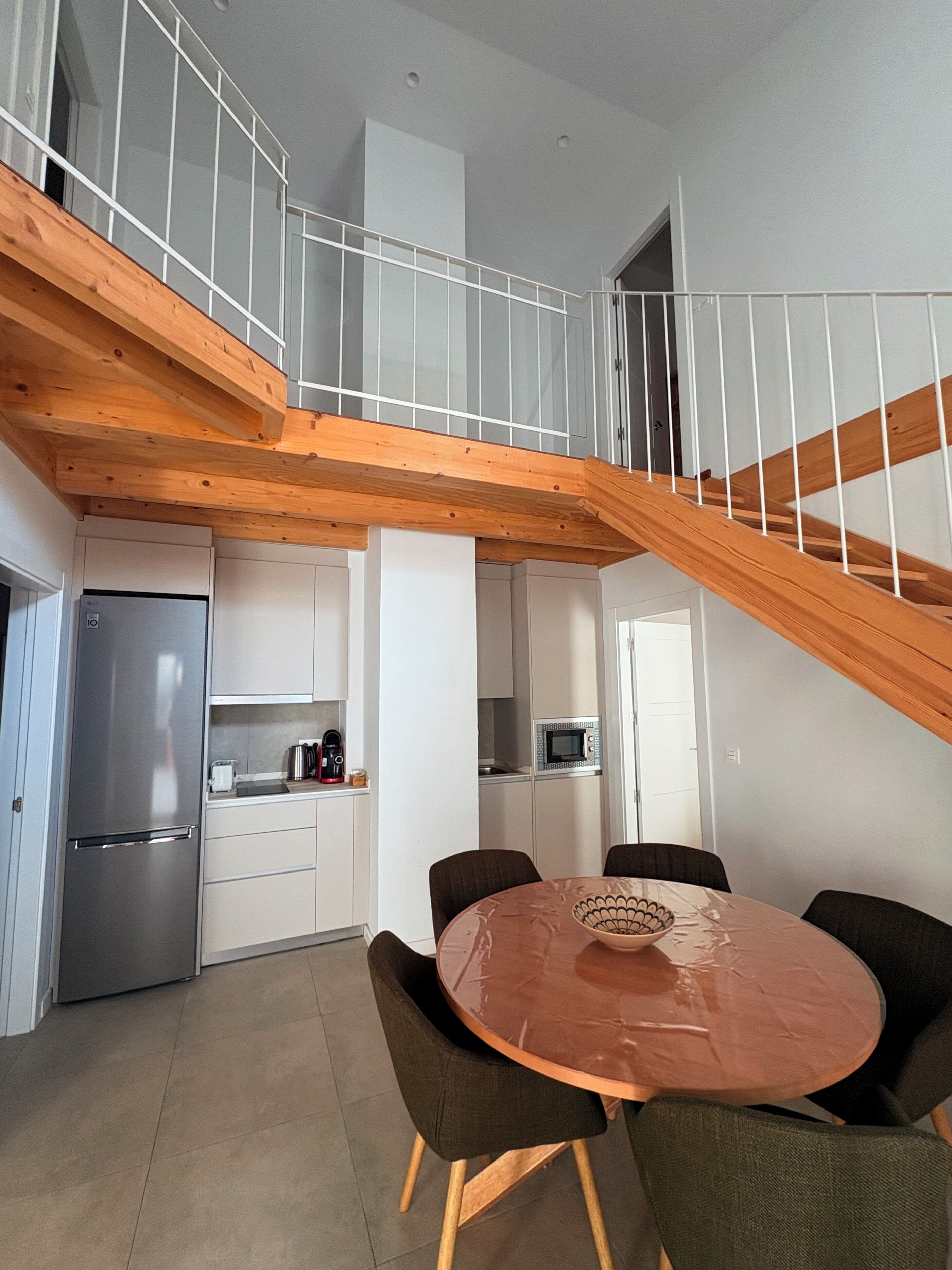 A modern kitchen with a central island, a dining area with a round table, and a wooden staircase.