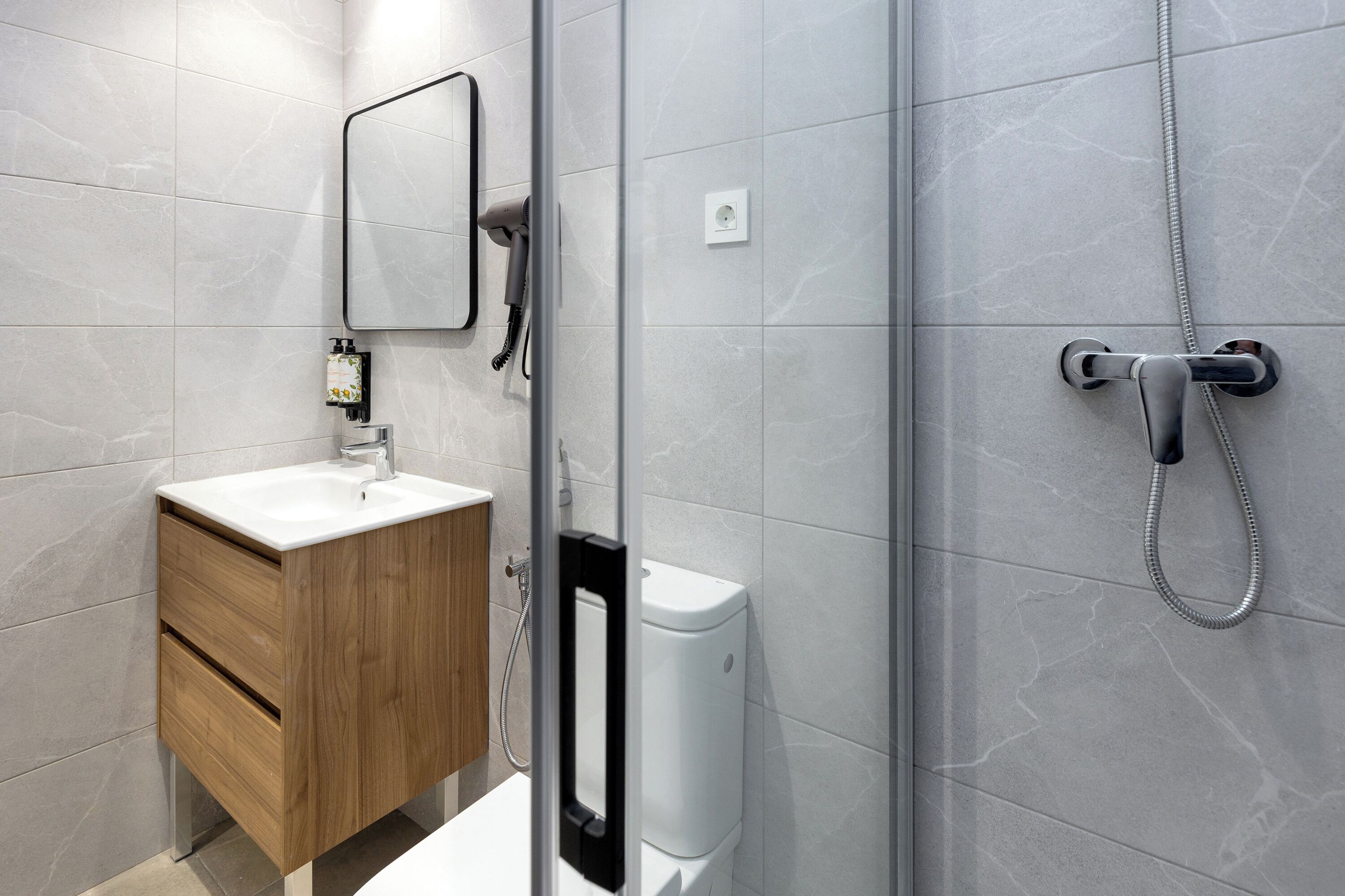 A modern bathroom with a wooden vanity, a white sink, a mirror, and a shower with a handheld showerhead.