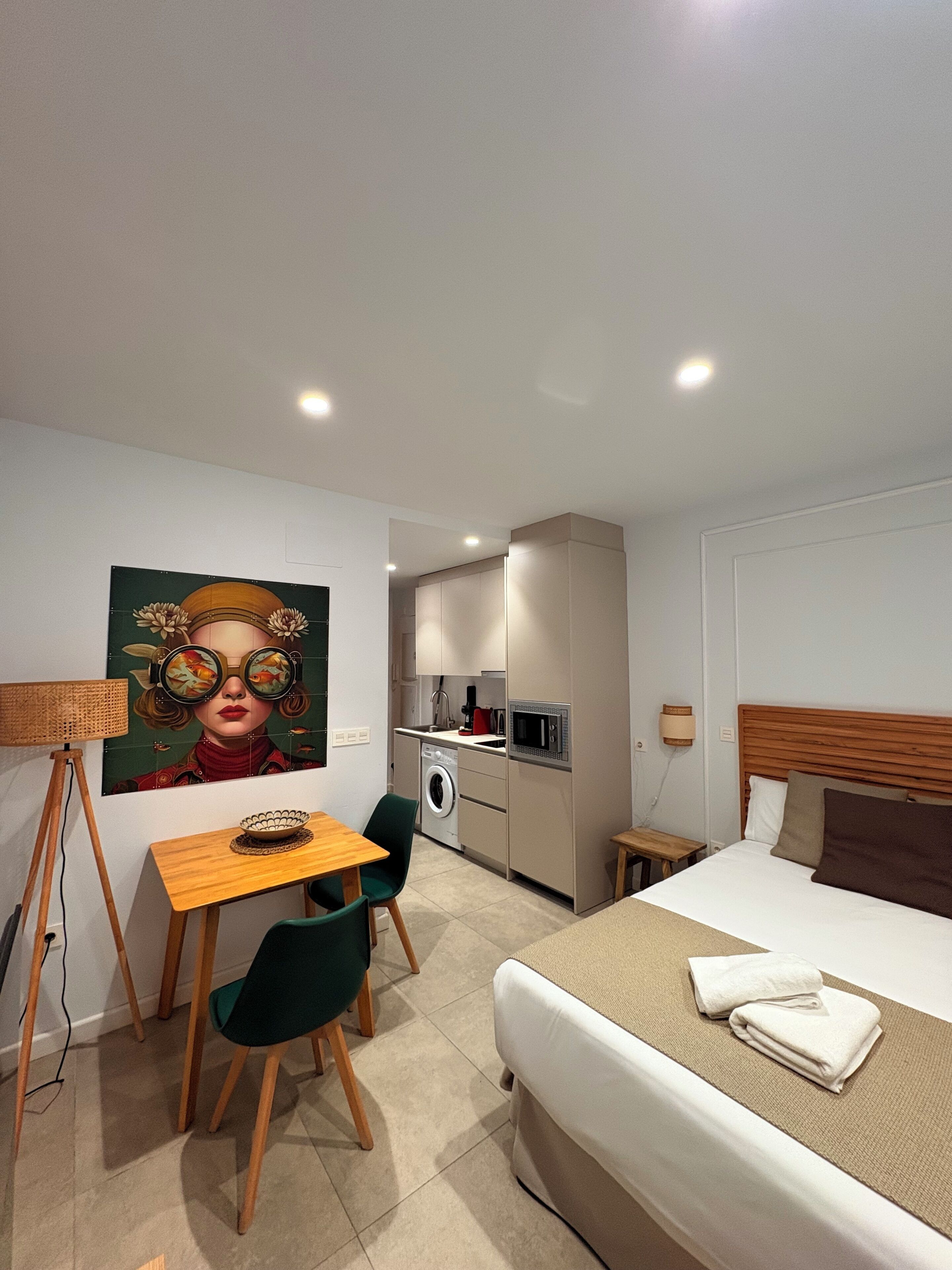 Una habitación de hotel moderna con cama, una pequeña mesa con sillas, una kitchenette y un cuadro en la pared.
