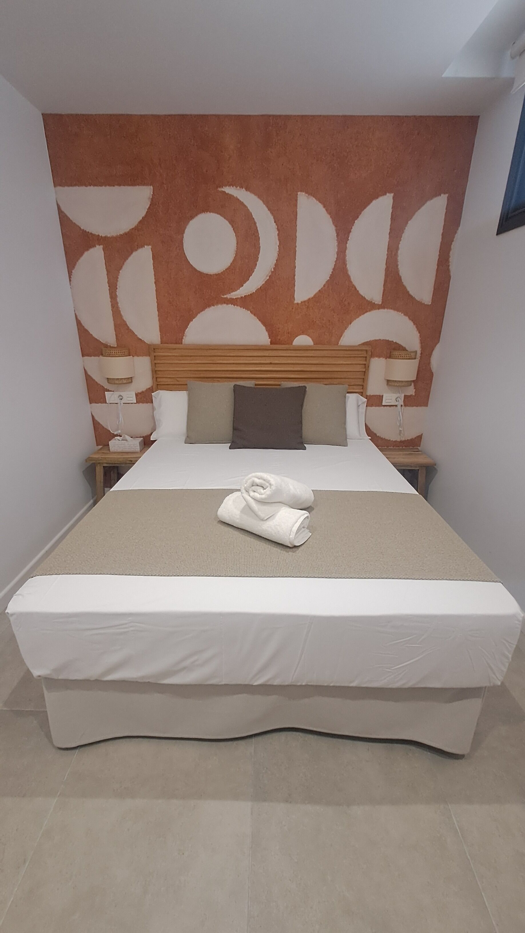 Una cama bien tendida con un edredón blanco y una toalla enrollada, un cabecero de madera y una pared con un diseño circular.