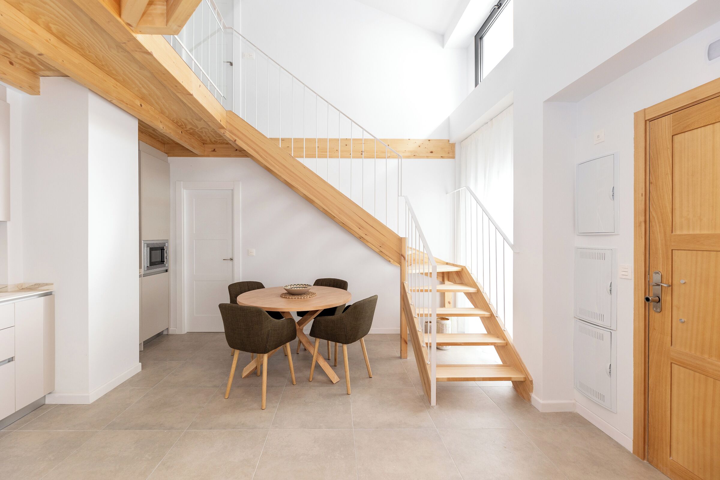 A modern kitchen with a dining area, wooden staircase, and white cabinetry.