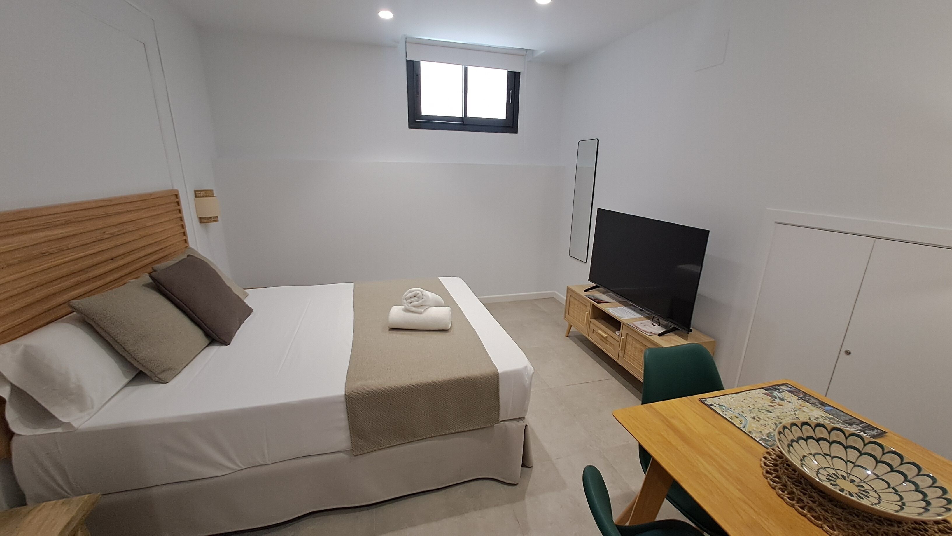 Una habitación pequeña y bien iluminada con una cama, un televisor, una mesa de madera y un ventanal.