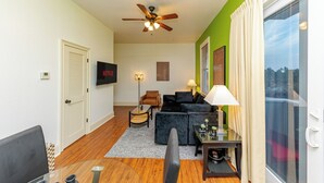 A living room with a ceiling fan, a black sofa, a glass coffee table, and a TV on the wall. - Modern King bed Haven/Free parking, Pool, Balcony (Santa Rosa Beach)