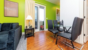 A modern living room with a green wall, a black sofa, a glass dining table with chairs, and a sliding glass door leading to a balcony. - Modern King bed Haven/Free parking, Pool, Balcony (Santa Rosa Beach)