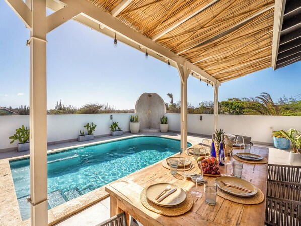 Un espace repas aménagé au bord de la piscine, avec une table pour deux, entouré de plantes en pot et offrant une vue dégagée sur la piscine.