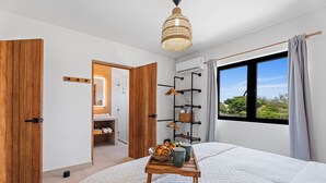 A modern bedroom with a large bed, a wooden bedside table with a tray of pastries, a hanging rack with clothes, and a window with a view of greenery.