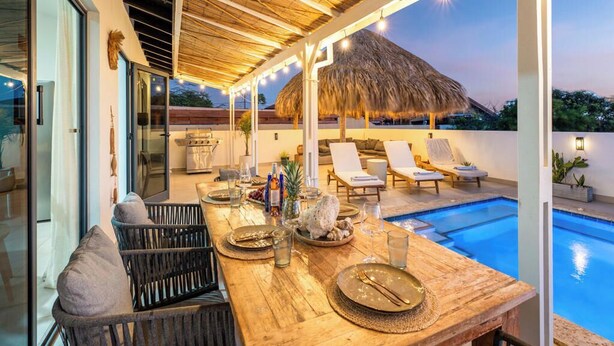 A dining area with a pool, lounge chairs, and a thatched roof structure.