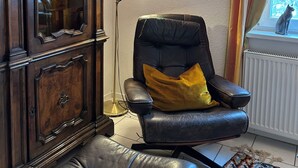 A room with a dark wooden cabinet, a black leather chair with a yellow pillow, a floor lamp, a framed botanical print on the wall, and a window with curtains.