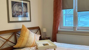 A neatly made bed with a tray of items, a framed picture on the wall, a window with curtains, and a radiator.