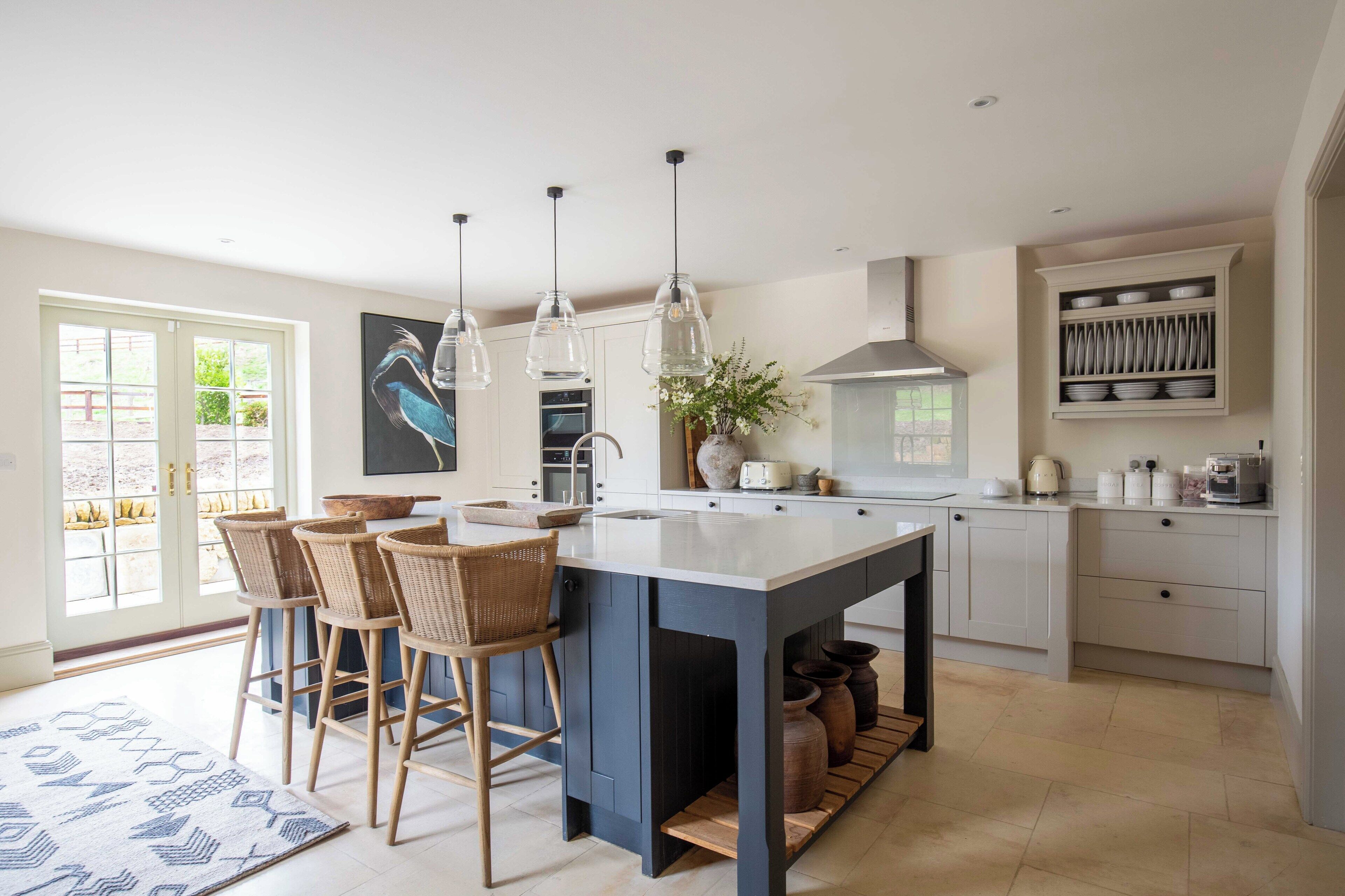 A modern kitchen with a central island, bar stools, pendant lights, and a view of the outdoors through large windows.