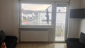 A living room with two black sofas, a wooden floor, a radiator, a large window with a view of a street and houses, and a TV mounted on the wall. - NR 3 Apartment on the upper floor 3 ZKB for companies/families (Vellmar)