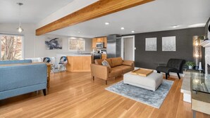 Ein geräumiger Wohnbereich mit Holzbalkendecke, einer Küche im Hintergrund und einem Essbereich mit blauem Sofa und weißem Hocker.