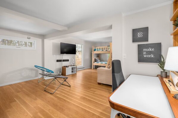 A modern living room with a wooden floor, a TV mounted on the wall, a bunk bed, and a desk with a lamp.