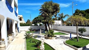 A courtyard with a pathway, palm trees, and a building with blue accents. - Villa Lea - Apartments Torre dell'Orso (Torre dell'Orso)