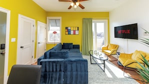 A living room with a sectional sofa, yellow walls, a ceiling fan, and a TV mounted on the wall. - Modern King bed Haven/Free parking, Pool, Balcony (Santa Rosa Beach)