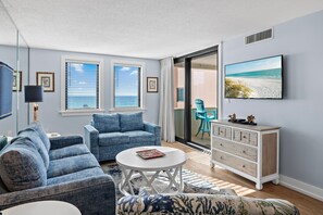 Ein Wohnzimmer mit einer Couch, Sesseln, einem Couchtisch, einem Fernseher und Blick auf den Strand durch die Glastür.