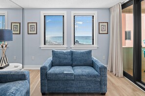 Un salon avec un canapé bleu, deux fenêtres équipées de stores, une lampe et une vue sur la plage.