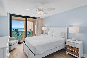 A hotel room with a bed, two bedside tables with lamps, a ceiling fan, a view of the ocean through a sliding door, and a balcony with chairs.
