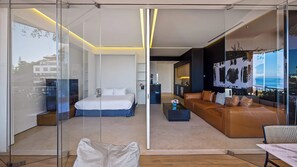 A modern hotel room with a glass partition, a bed, a sofa, and a view of the sea.