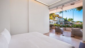 A modern hotel room with a large bed, a glass wall, and a balcony with outdoor seating.