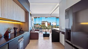 A modern kitchen with a built-in oven and a large refrigerator. There is a dining area with a sofa and a table, and a balcony with a view of the city.