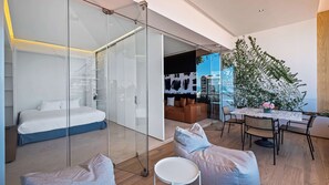 A modern bedroom with a glass partition, a bed, a small round table, and a seating area with cushions.